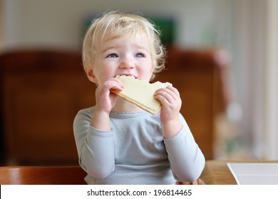 Funny Little Child, Blonde Toddler Girl, Sitting On High Feeding Chair In The Kitchen Eating Sandwich, Bread With Butter And Cheese, For Breakfast On A Sunny Morning