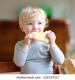 Funny Little Child, Blonde Toddler Girl, Sitting On High Feeding Chair In The Kitchen Eating Sandwich, Bread With Butter And Cheese, For Breakfast On A Sunny Morning