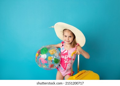 Funny little child girl with beach ball holding yellow suitcase above blue background. Summer and vacation concept.