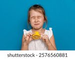 Funny little child girl try to taste fresh lemon, wrinkles and makes a grimace. Sour taste, facial emotions negative, in white dress, isolated blue background, copy space. High quality photo