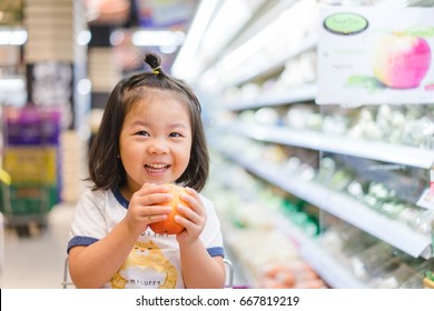 Funny Little Child Smile And Holding Apple.Asian Kid Shopping In Supermarket.Little Asian Toddler Girl Sitting In The Trolley During Family Shopping In Hypermarket.