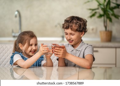 Funny Little Children Drink Water In The Kitchen At Home. Boy And Girl Are Smiling Happily, Holding A Glass Of Clean Water In Their Hands And Looking At Them. Water Balance. Prevention Of Dehydration