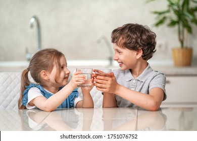 Funny Little Children Drink Water In The Kitchen At Home. The Boy And Girl Are Smiling Happily, Holding A Glass Of Clear Water And Looking At Each Other. Water Balance. Prevention Of Dehydration