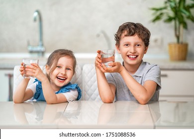 Funny Little Children Drink Water In The Kitchen At Home. The Boy And Girl Are Smiling Happily, Holding A Glass Of Clean Water And Want To Drink Water. Water Balance. Prevention Of Dehydration.