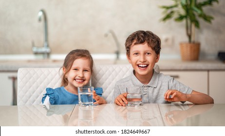 Funny Little Children Drink Water In The Kitchen At Home. The Boy And Girl Are Smiling Happily, Holding A Glass Of Clean Water And Want To Drink Water. Water Balance. Prevention Of Dehydration.