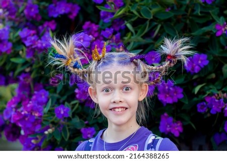 Funny little cute caucasian girl with braids for crazy hair day at school on purple flowers tibouchina outside nature background