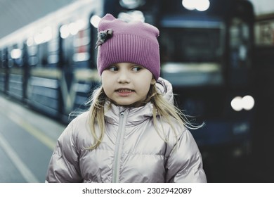 Funny little girl 5 year old posing on platform underground metro station, looking away. Adorable kid small girl passenger waiting train in subway. Public urban transport concept. Copy ad text space