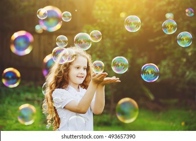 Funny little girl catching soap bubbles in the summer on nature. Background toning instagram filter. Happy childhood concept.