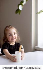 Funny Little Girl With Dirty Face Eating Plain Homemade Yogurt From Glass Jar Using Spoon. Vertical. 18-24 Months Kid Girl In Black T-shirt Eating Yogurt And Looking Window. Vertical Format