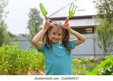 Funny Little Girl Holding Small Gardening Tools , Standing In The Garden On A Sunny Day