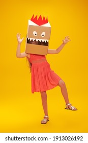 Funny Little Girl Playing With A Cardboard Dragon, Dinosaur. Childhood Dreams. Fantasy, Imagination. Studio Portrait On A Yellow Background.