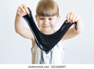 Funny Little Girl Playing With Slippery Slime. A Cheerful Little Girl Holds In Her Hands And Stretches The Black Slime. A Happy Child Is Playing With A Slippery, Sticky Toy. Entertainment For Children