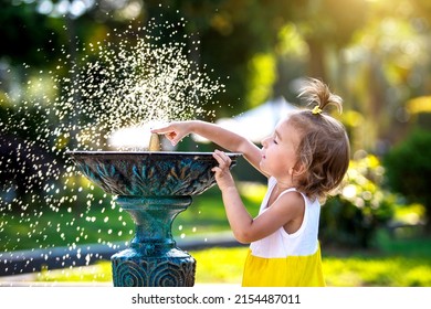 A Funny Little Girl Is Playing With The Spray Of A Drinking Water Fountain In The Park. Childhood, Child, Summer Time, Heat. Solar Protection For Children's Skin,