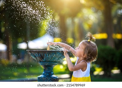 A Funny Little Girl Is Playing With The Spray Of A Drinking Water Fountain In The Park. Childhood, Child, Summer Time, Heat. Solar Protection For Children's Skin,