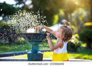 A funny little girl is playing with the spray of a drinking water fountain in the park. Childhood, child, summer time, heat. Solar protection for children's skin,