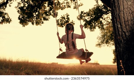 Funny little girl plays swings under old tree at sunset time. Happy little girl enjoys flight feeling on swings in country field on summer holiday. Little girl rides handmade swing in country garden