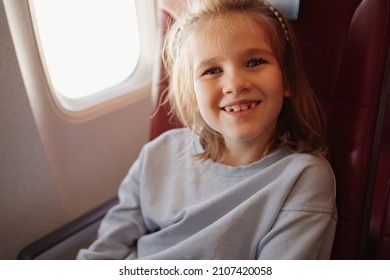 A Funny Little Girl Sits In An Airplane Seat By The Window. Flights With Children In Economy Class. Sale Of Vouchers And Plane Tickets.