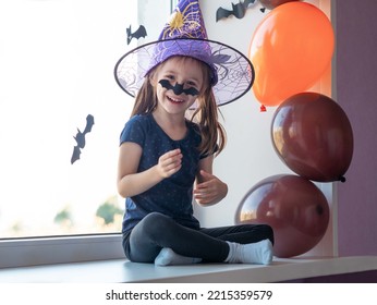 Funny Little Girl Smiling In A Witch Hat, Stuck A Bat On Her Nose, Laughing. Happy Halloween.