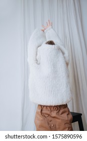 Funny Little Girl Struggling To Put On White Fluffy Knitted Sweater. Her Head And Arms Still Inside. She's Squeezing Hands Out Of Sleeves. At Home, In Front Of A Curtain. Half Length.