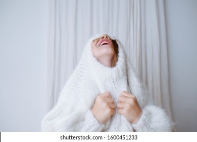 Funny Little Girl Struggling To Put On White Fluffy Knitted Sweater. She's Squeezing Her Head Out Of The Collar, Pulling Fabric With Hands. At Home, In Front Of A Curtain. Half Length.