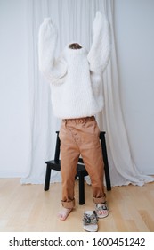 Funny Little Girl Struggling To Put On White Fluffy Knitted Sweater. It Does Not Slide Down On Inself, So Her Head And Arms Still Inside. At Home, In Front Of A Curtain. Full Length.