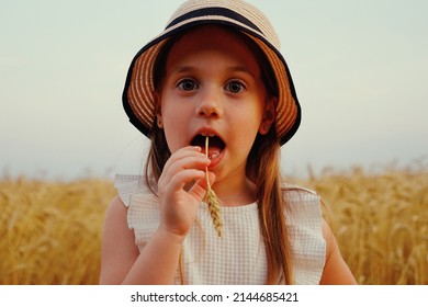 Funny little girl wearing white dress and straw hat standing in wheat field with spikelet in her mouth, smiling and looking at camera. Closeup happy kid in countryside. Concept of childhood
