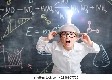 A funny little girl in white shirt and glasses stands with a strange invention on her head against the blackboard with chemical formulas and diagrams. Educational concept. 