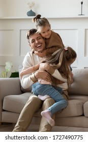 Funny Little Girls Hugging Cuddling Smiling Laughing Father Sitting On Comfortable Couch In Living Room, Excited Father Having Fun With Two Adorable Little Daughters, Enjoying Free Time Together