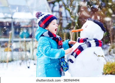 Funny little kid boy making a snowman and eating carrot. child playing and having fun with snow on cold day. Active outdoors leisure with kids in winter.