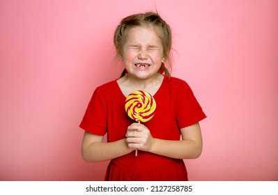 Funny Little Kid Close Eyes With Candy In Hands. Child With Missing Milk Tooth. Little Girl Licking A Lollipop Isolated On Pink Background. Dental Health.