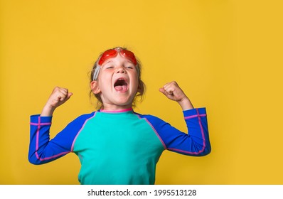 Funny Little Kid Girl Shouting With Hands Up Isolated On Yellow Background. Strong And Healthy Child Can Swim In Water Park. Portrait Of Small Child In Pink Goggles And Blue Swimsuit Ready To Dive