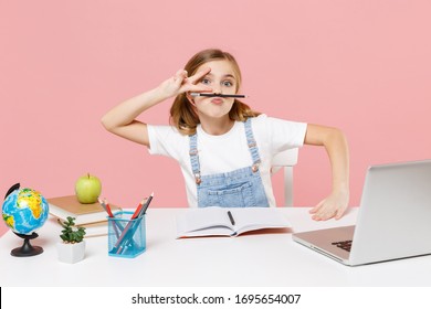 Funny little kid schoolgirl 12-13 years old sit study at white desk with pc laptop isolated on pink background. School distance education at home concept. Hold pencil with lips, showing victory sign