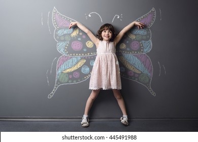 Funny little kid smiles with rised hands with the dream of being the butterfly. Huge depicted with chalk insect on the neutral background.