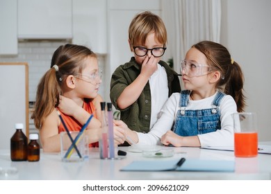 Funny Little Kids Doing Home Science Project, Looking At Each Other In Uncertainty, Not Sure What To Do. All Behid Table, Wearing Glasses. Chemical Glassware And Colored Liquids On The Table.