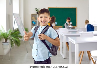 Funny little schoolboy showing thumbs up showing positive approval of great choice. Portrait of cheerful first grader with backpack on his shoulders standing in classroom and smiling at camera. - Powered by Shutterstock