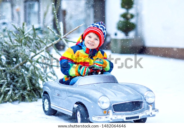 Funny little smiling\
kid boy driving toy car with Christmas tree. Happy child in winter\
fashion clothes bringing hewed xmas tree from snowy forest. Family,\
tradition, holiday.