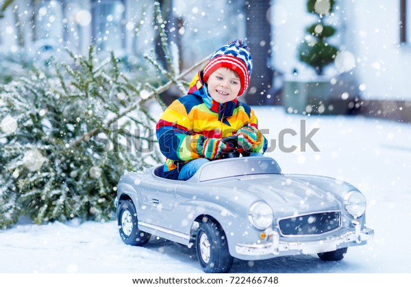 Funny little smiling\
kid boy driving toy car with Christmas tree. Happy child in winter\
fashion clothes bringing hewed xmas tree from snowy forest. Family,\
tradition, holiday