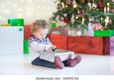 Funny little toddler girl opening her Christmas present under a beautiful Christmas tree