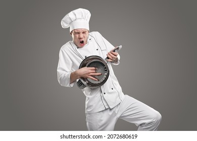 Funny male cook in white uniform pretending being crazy guitarist playing colander impromptu guitar in studio against gray background