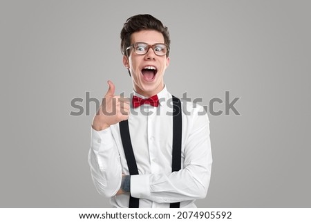 Funny male geek in glasses showing like sign and looking at camera with opened mouth on gray background