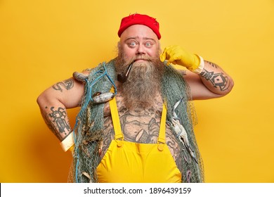 Funny male sailor curls mustache has smoking pipe in mouth wears red hat overalls and gloves poses against vivid yellow background thinks about sea adventure looks with widely opened eyes at camera