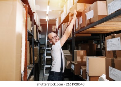 Funny male warehouse worker reaching up, putting box on a shelf, standing between high rows of shelves with boxes. Smiling at the camera - Shutterstock ID 2161928543