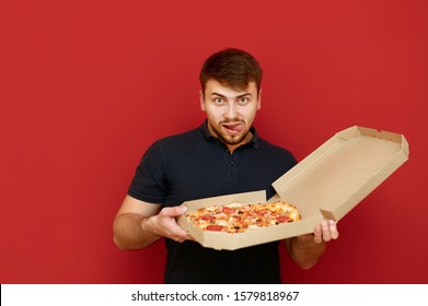 A funny man with a beard holds a pizza box in delivery and looks into the camera with a hungry lustful face. Portrait of hungry man with pizza box on red background. Fast junk food concept.