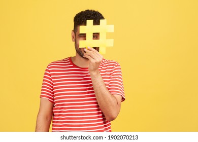 Funny man blogger or smm manager in striped t-shirt hiding face behind yellow hashtag sign, teaching to tag topics on websites, giving recommendations. Indoor studio shot isolated on yellow background