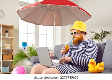 Funny man in classic bathing suit enjoying summer vacation at home. Eccentric bearded man in sunglasses and panama hat sitting under umbrella, drinking cocktail and using laptop in living room