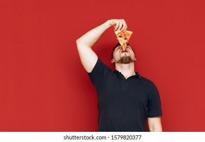 Funny man in dark t-shirt eating pizza on red background, lifting his head up and thrusting a piece into his mouth. Photo of man eating the last slice of pizza, isolated. Fast food. Copy space