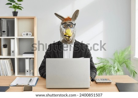 Funny man in donkey head mask sitting at desk in office. Stupid strange male office worker, employee in formal suit and glasses using laptop computer and looking at camera