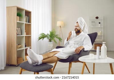 Funny man drinking coffee in morning and enjoying beauty treatments and luxury relaxation at home spa. Man with towel on his head sits on armchair in white bathrobe and with cosmetic mask on his face. - Powered by Shutterstock