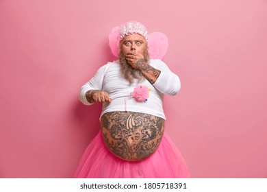 Funny Man In Fairy Costume Has Shocked Reaction On Something Terrible, Nurses Children Alone Without Wife, Stares With Bugged Eyes, Shows Fat Belly, Holds Magic Wand, Isolated On Pink Studio Wall