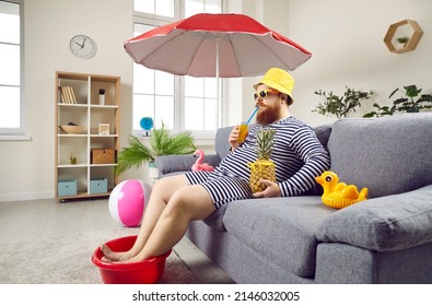 Funny man in living room at home imagines that he is resting on sea and sunbathing on beach. Chubby man wets his feet in plastic bowl and drinks cocktail while sitting on sofa under beach umbrella. - Shutterstock ID 2146032005